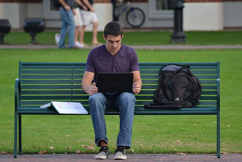 Student working on a laptop outdoors with an open book beside them